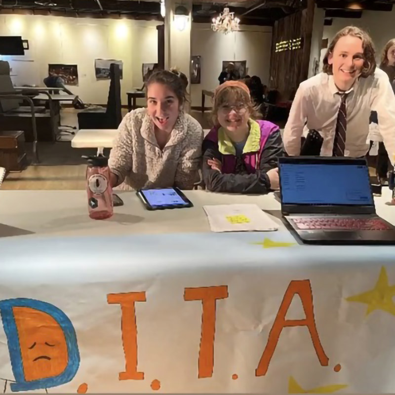 Three students sitting behind a table with a banner reading D.I.T.A. and a laptop.