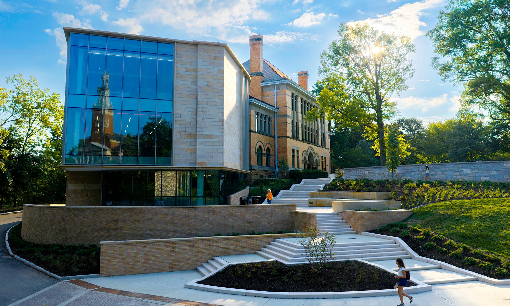 Modern building with large glass windows, brick facade, multi-level steps, landscaping, and a person walking in the foreground on a sunny day.