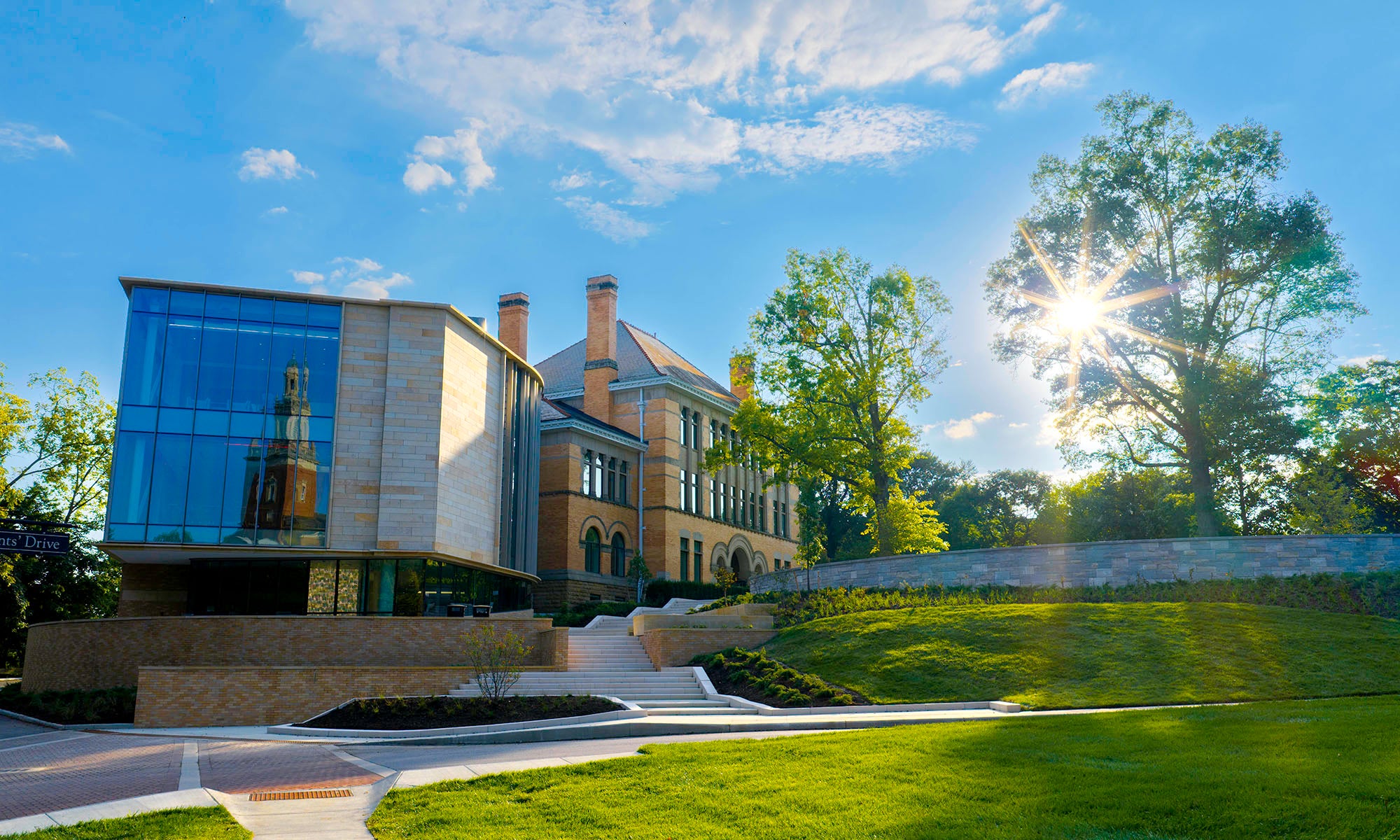 Modern glass extension and historic brick building surrounded by green lawn and trees under a bright, sunny sky.