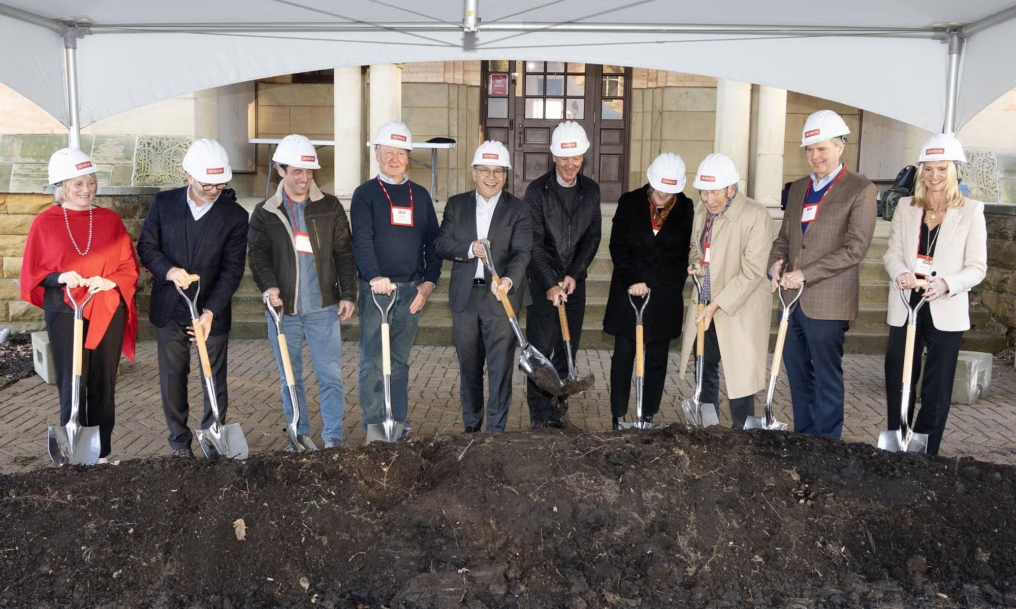 Ten people in hard hats stand in a row, holding shovels and turning soil at a groundbreaking ceremony in front of a building.
