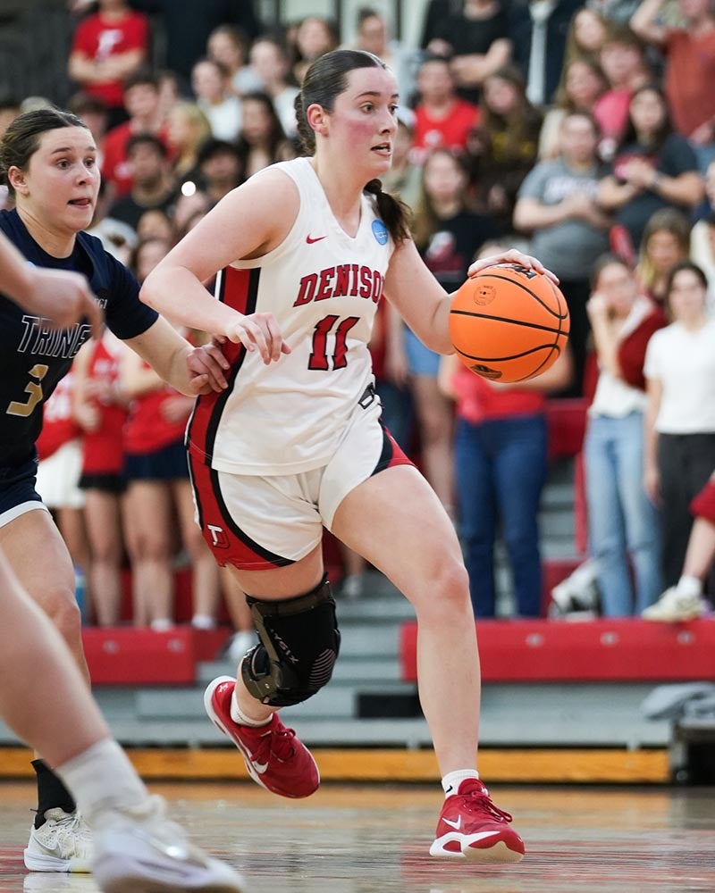 A basketball player in a white Denison uniform dribbles an orange basketball down the court during a game, with another player in a dark uniform nearby and a crowd in the background.