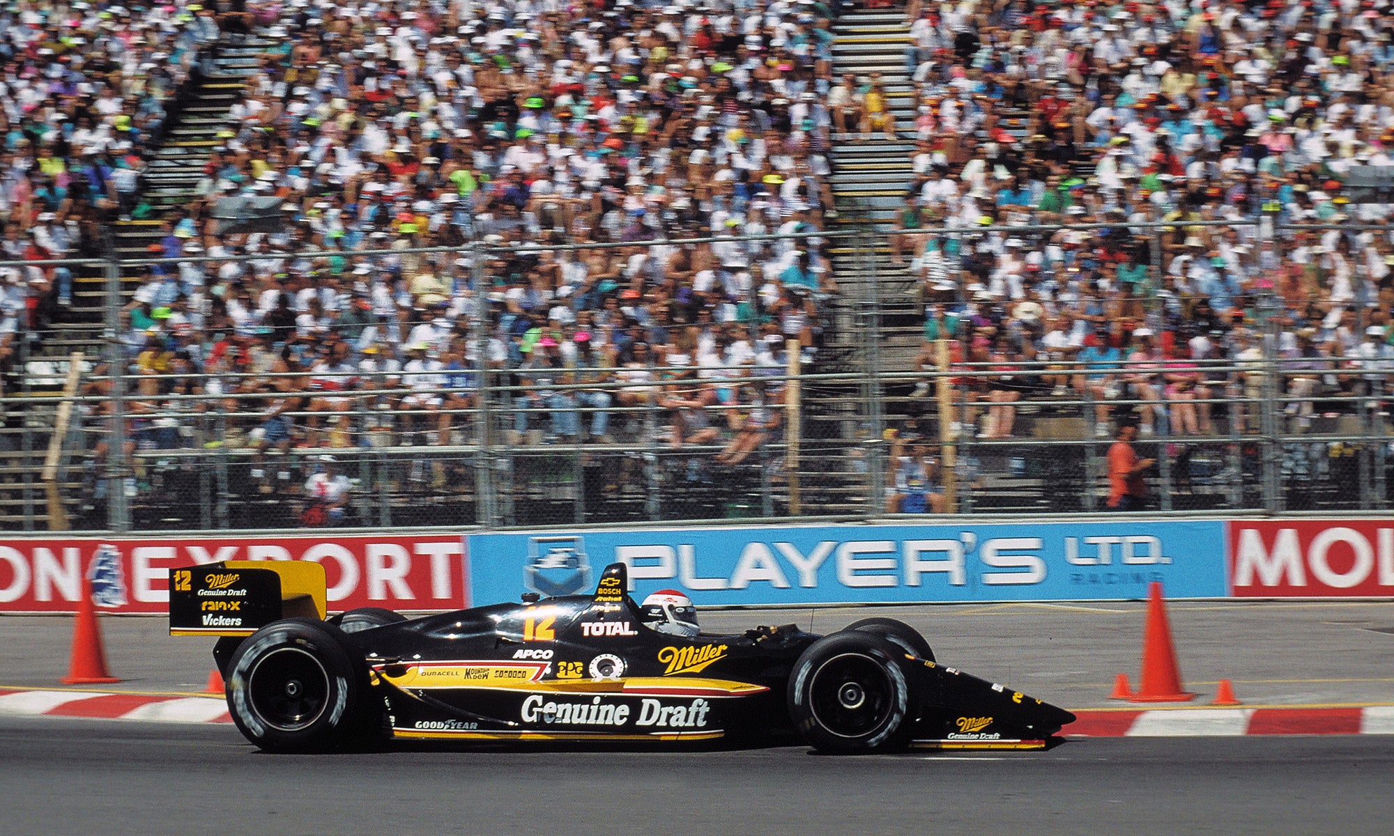 Black and yellow open-wheel race car on a track with a large crowd in the grandstands and barriers displaying sponsor logos in the background.