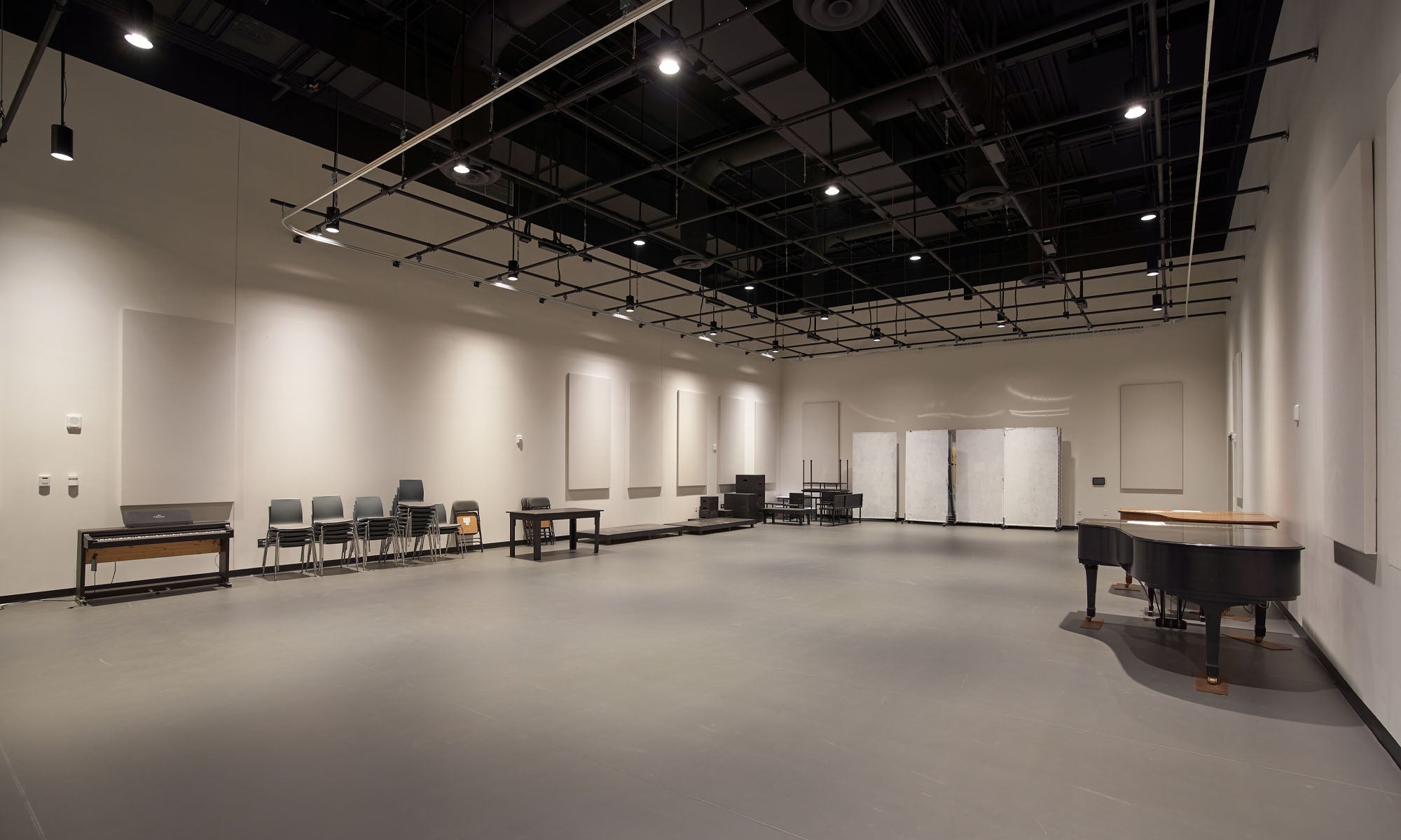A large performing arts rehearsal space with light-gray and white walls and floors, featuring a lighting grid hanging from a dark ceiling, piano, and furniture.