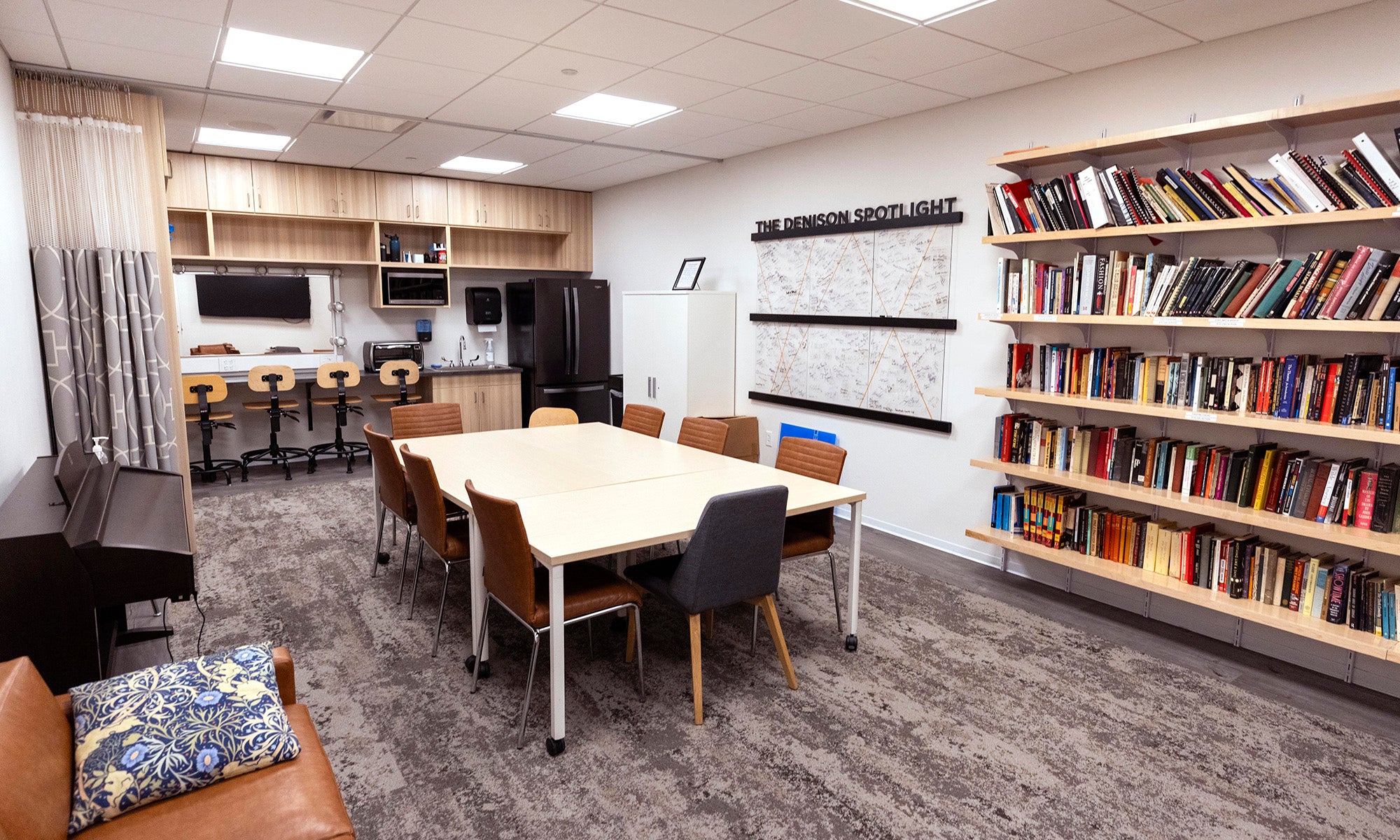 A modern office space with a conference table, chairs, bookshelves filled with books, a lounge area, and a kitchenette with stools and a refrigerator.