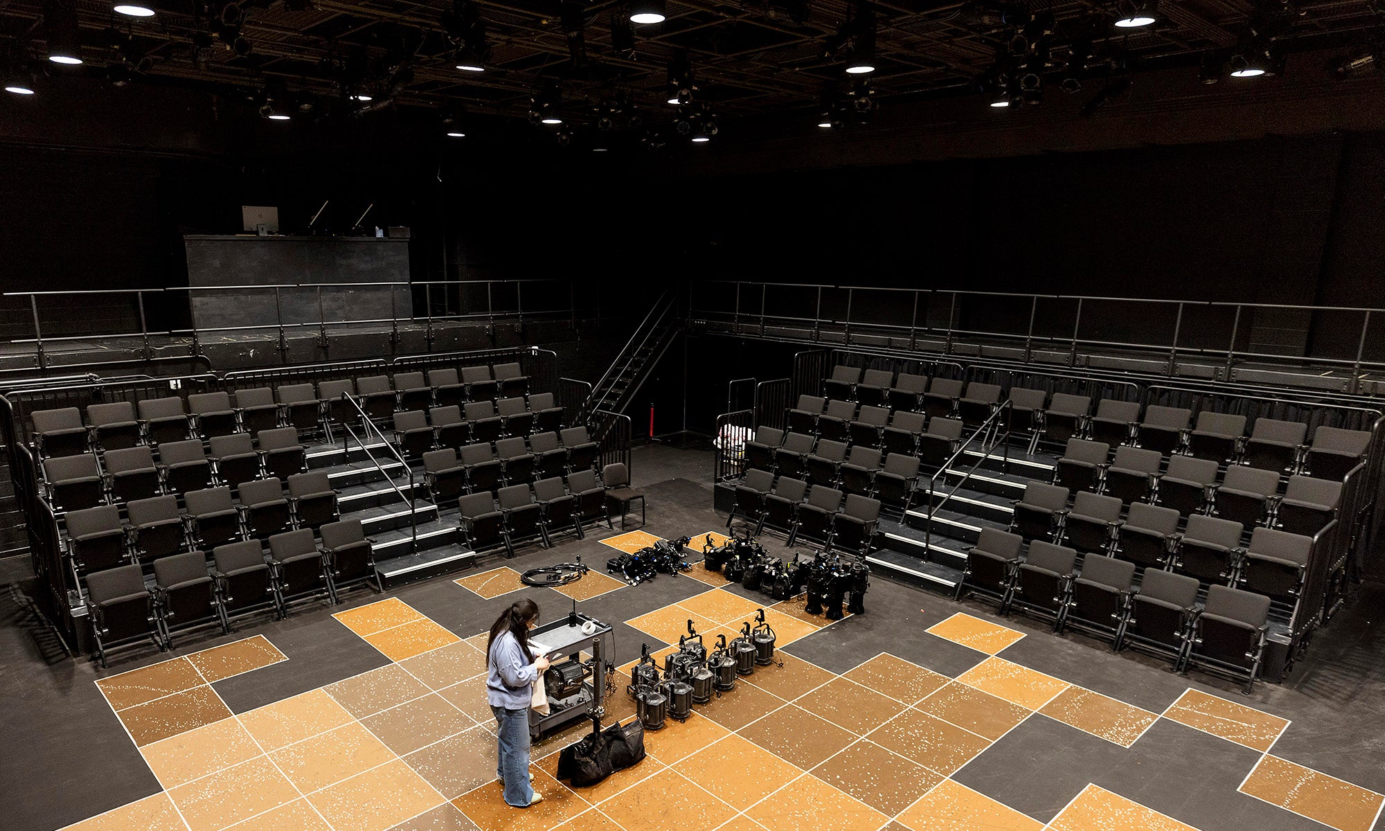 A person stands near equipment on a stage with patterned flooring in a black box theater; empty tiered seating surrounds the stage.