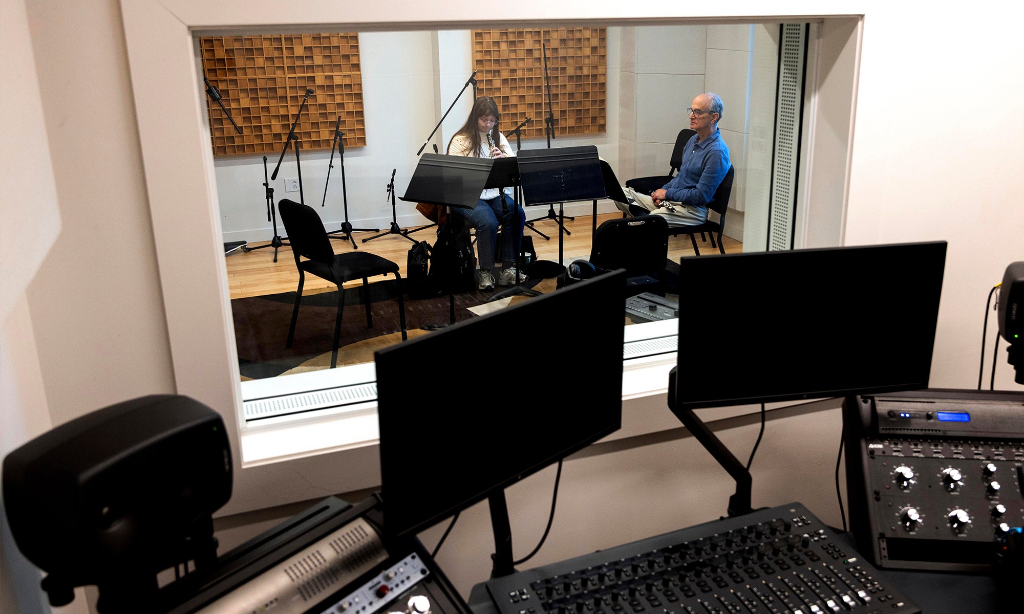 A recording studio window; recording equipment in the foreground, and two people sitting in the recording booth on the other side of the window.