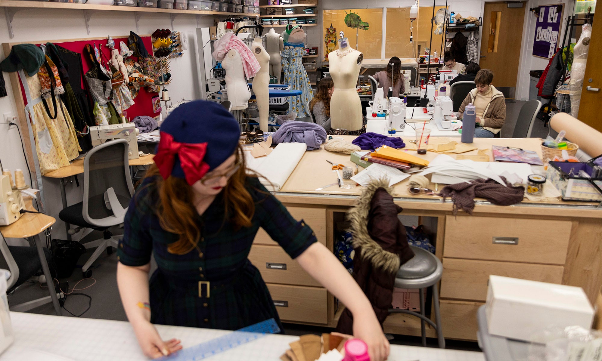 A person working in a theatre costume workshop filled with work tables, sewing machines, fabric, and mannequins.