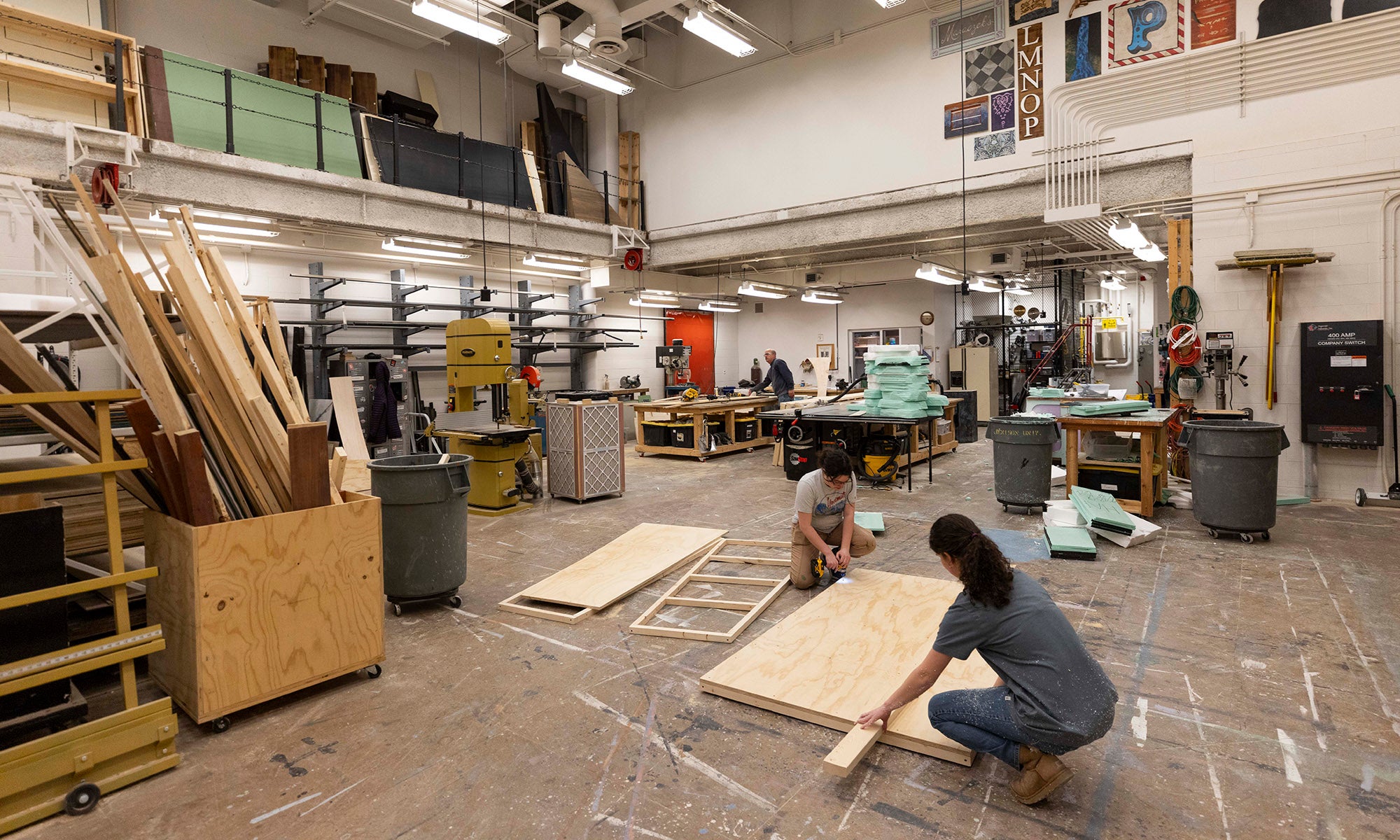 Two people working in a large scene shop filled with stacks of wood, tools, and ongoing theatre set construction.