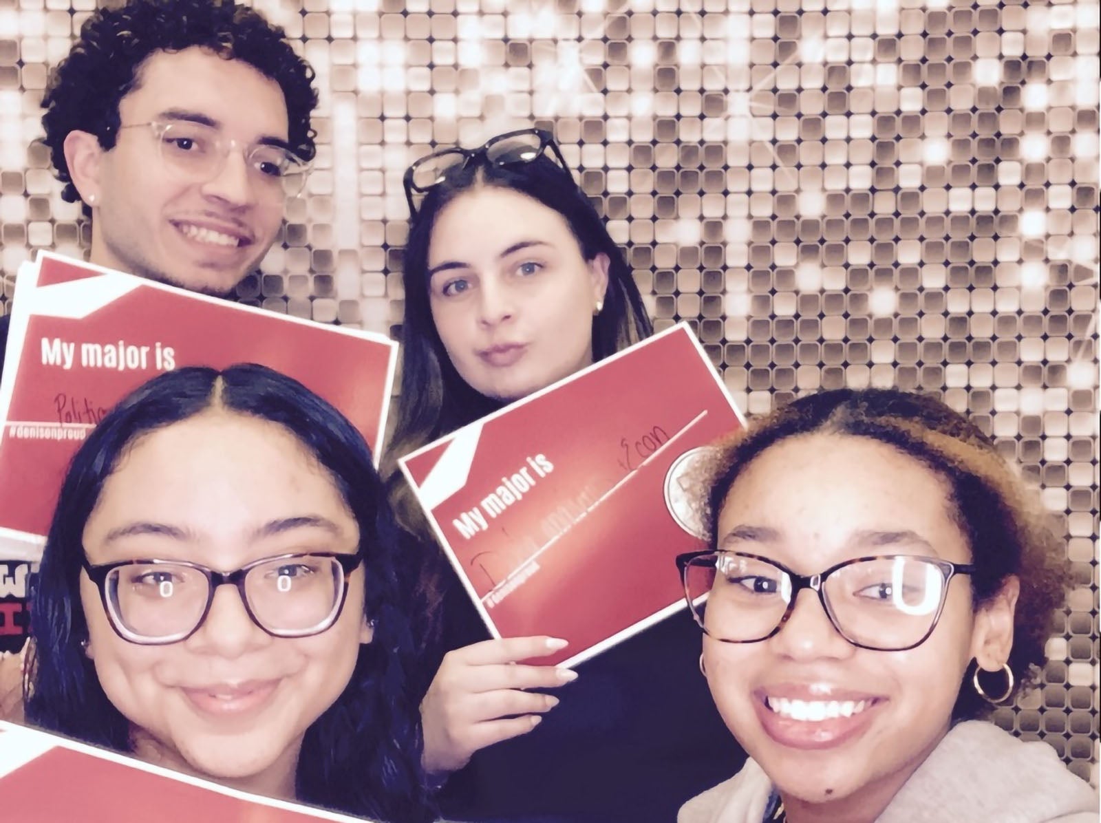 Four people pose in a photo booth holding red signs that say “My major is” with personal inscriptions, against a gold sequin backdrop.