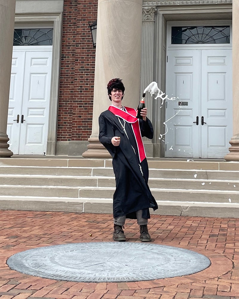 A person in a graduation gown and red stole stands outside in front of large columns and white doors, popping a bottle with liquid spraying out.
