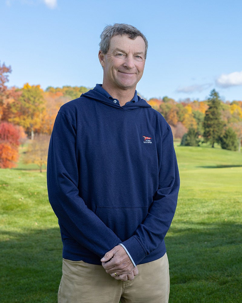 A person stands on a grassy field with colorful autumn trees and a blue sky in the background.