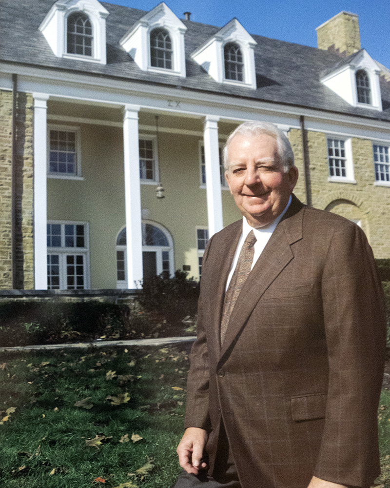 A man in a brown suit jacket standing in front of a large two-story house.