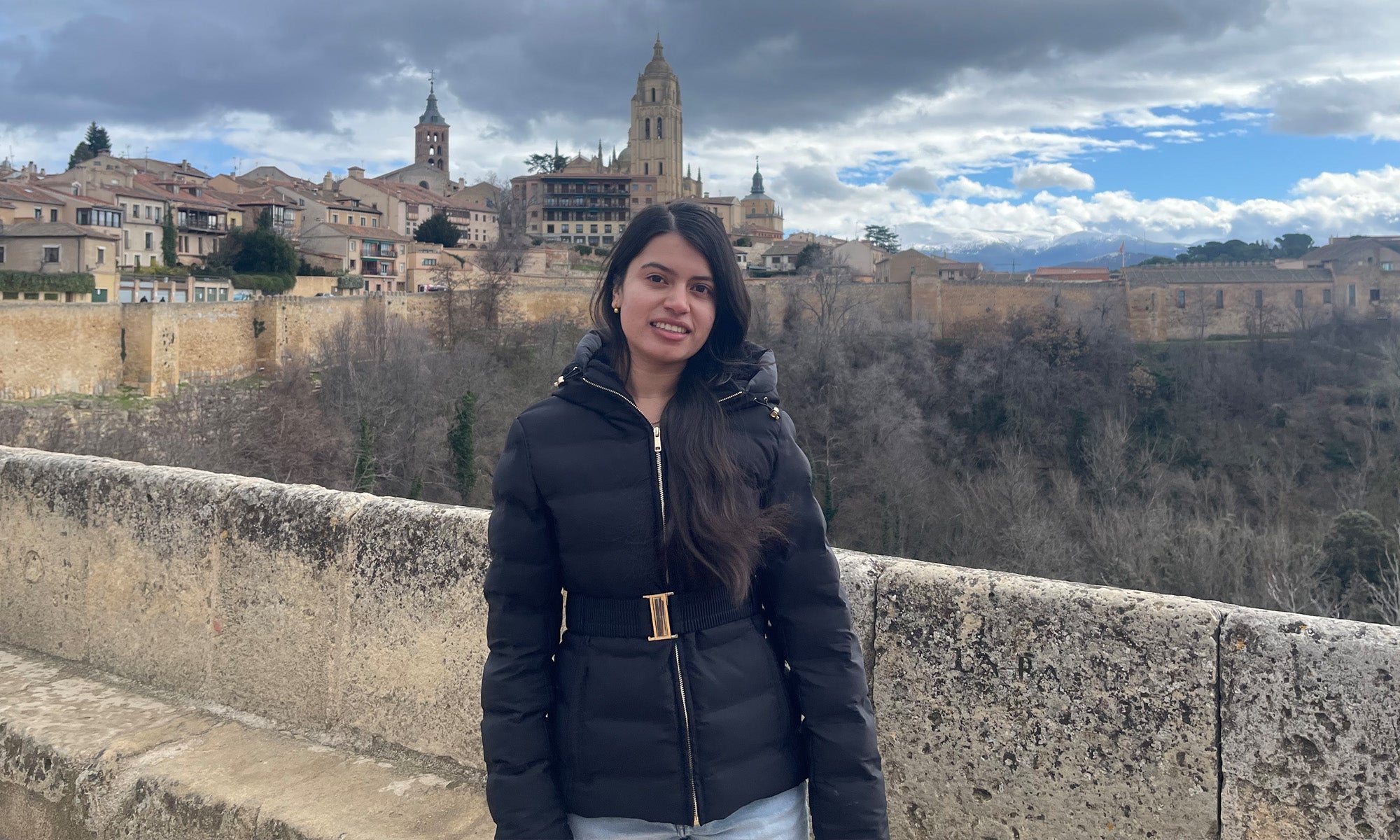 A woman stands on a stone bridge wearing a black jacket, with historic buildings and a church in the background under a cloudy sky, capturing the essence of study abroad adventures.