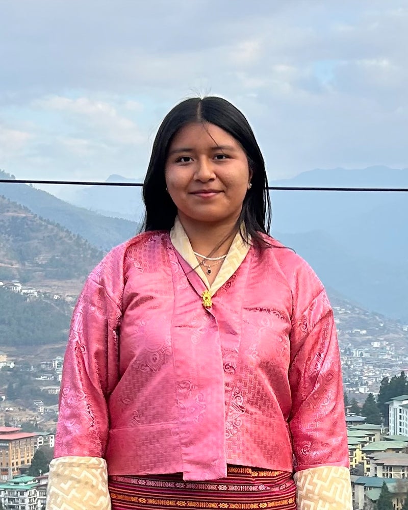 A study abroad student in a pink traditional outfit stands outdoors with a mountainous landscape and a city in the background.