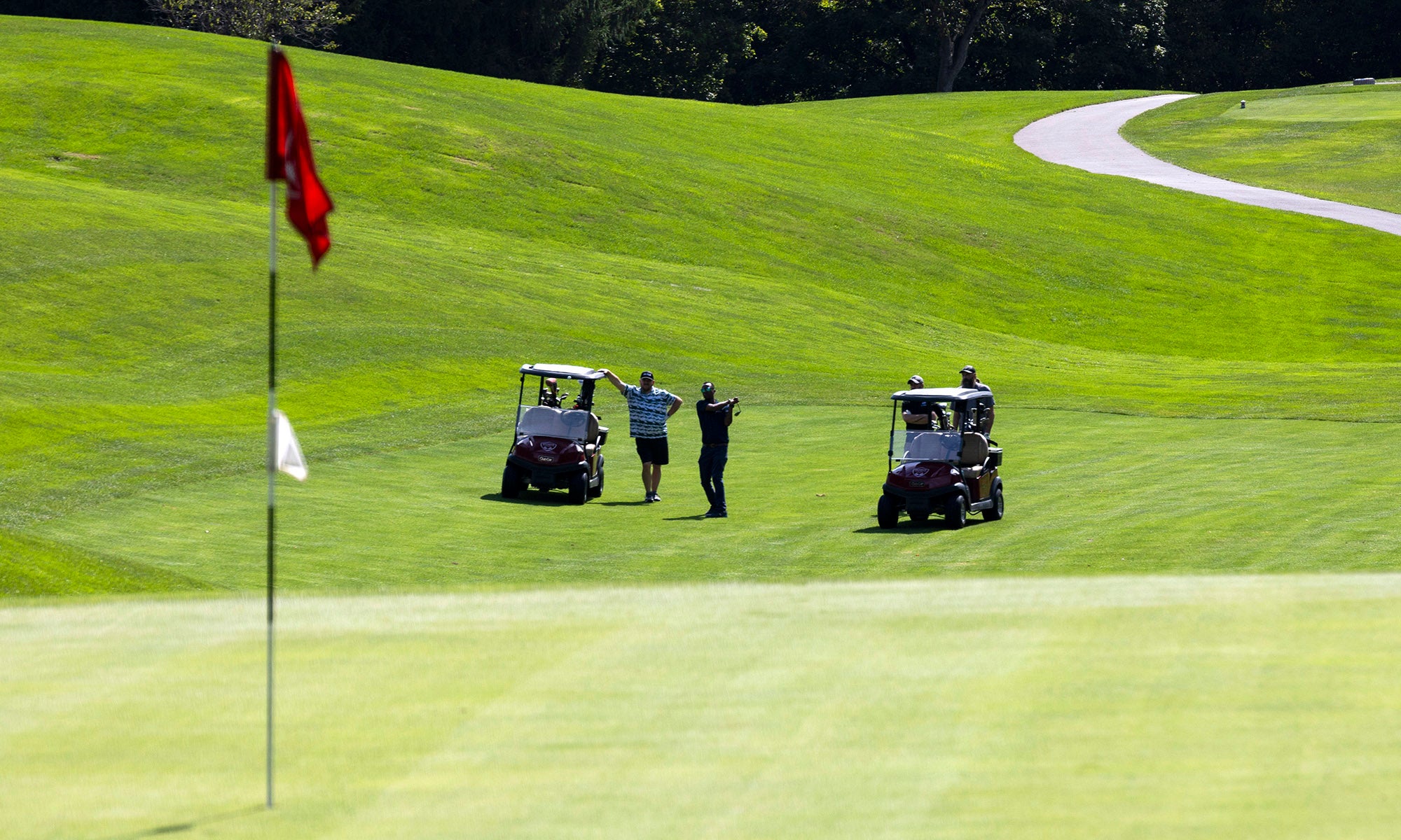 Two golf carts and three people stand on a grassy fairway near a putting green with a red flag, under clear daylight.