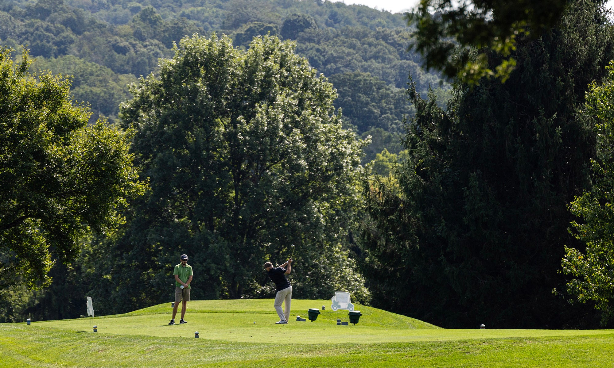 Two people stand on a golf course tee box surrounded by trees, with one person swinging a golf club and the other observing.