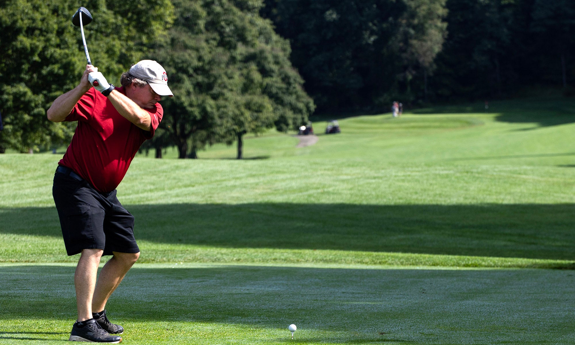 A person wearing a red shirt and black shorts is preparing to swing a golf club on a grassy golf course with trees in the background.