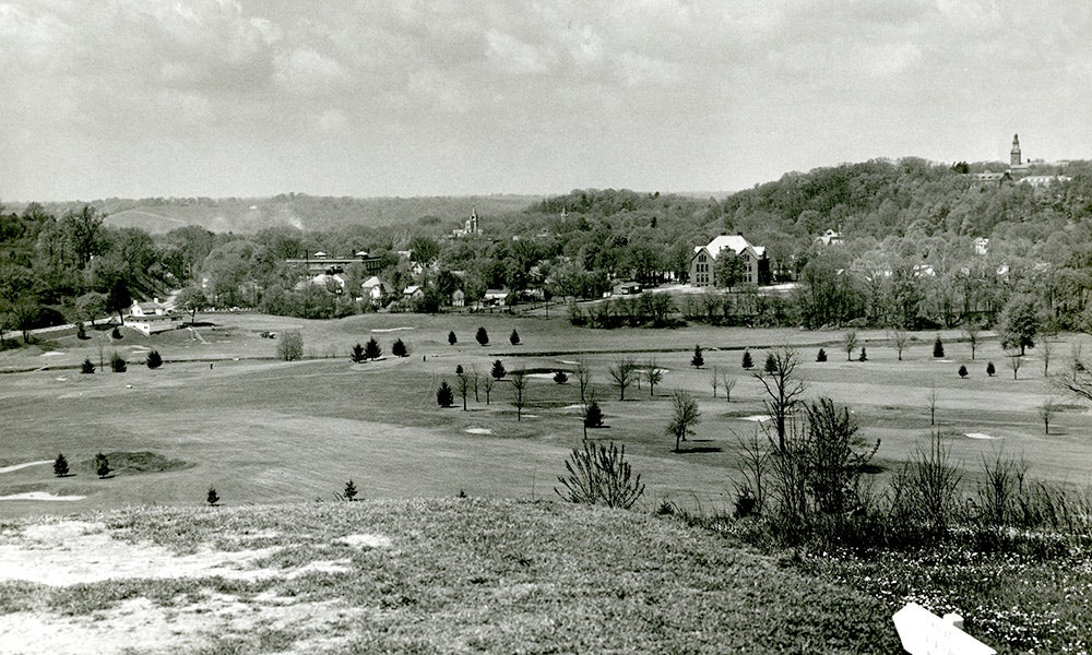 Black and white photo of a hilly landscape with scattered trees, open fields, and buildings in the distance under a cloudy sky.