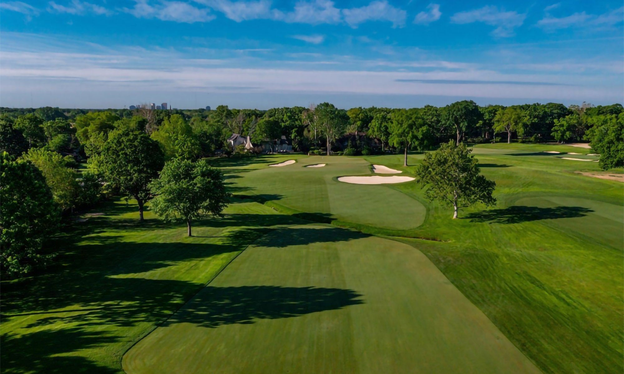 Aerial view of a green golf course with sand bunkers, trees, and a blue sky in the background.