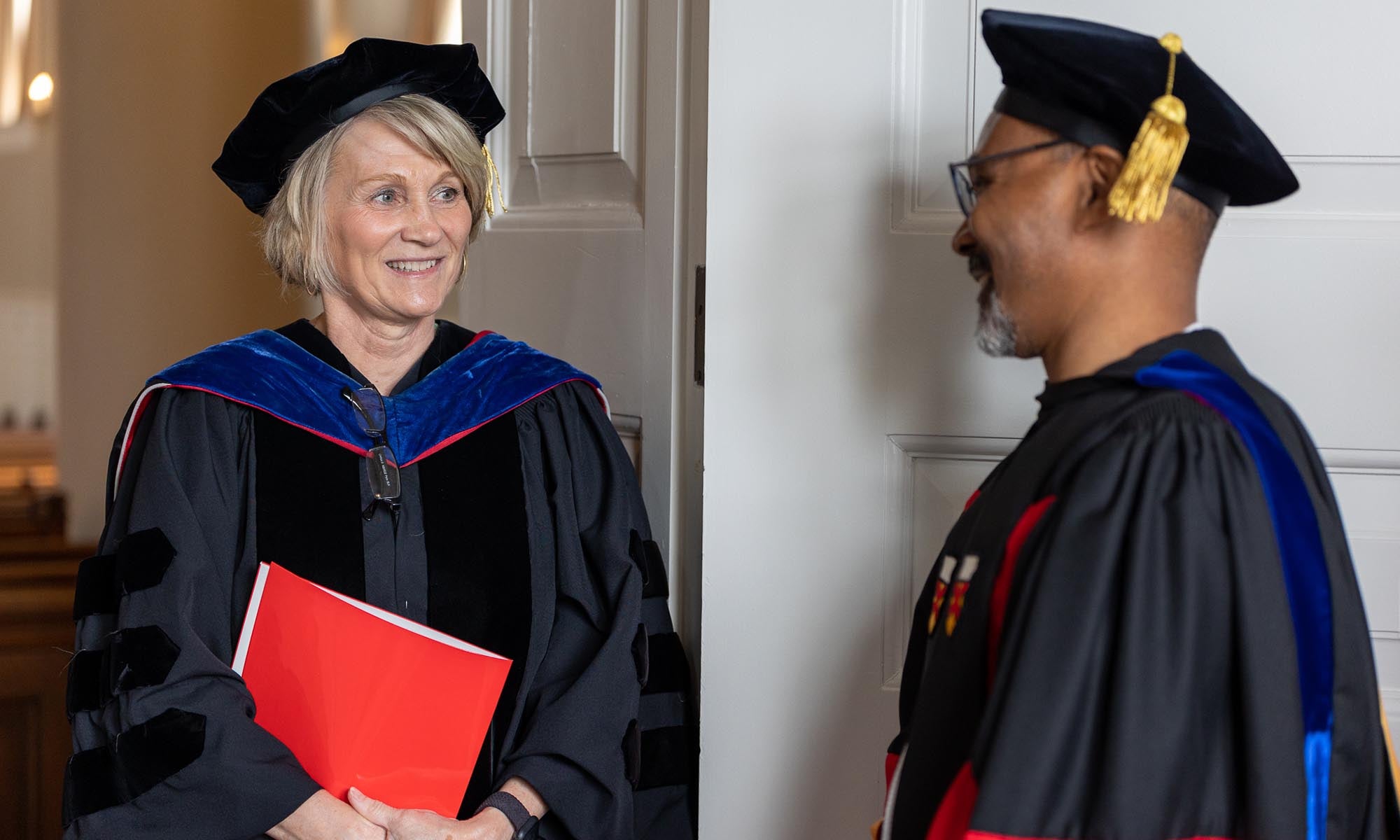 Two people wearing academic regalia and hats stand indoors, facing each other and smiling. One person, Denison Provost Kim Coplin, holds a red folder.