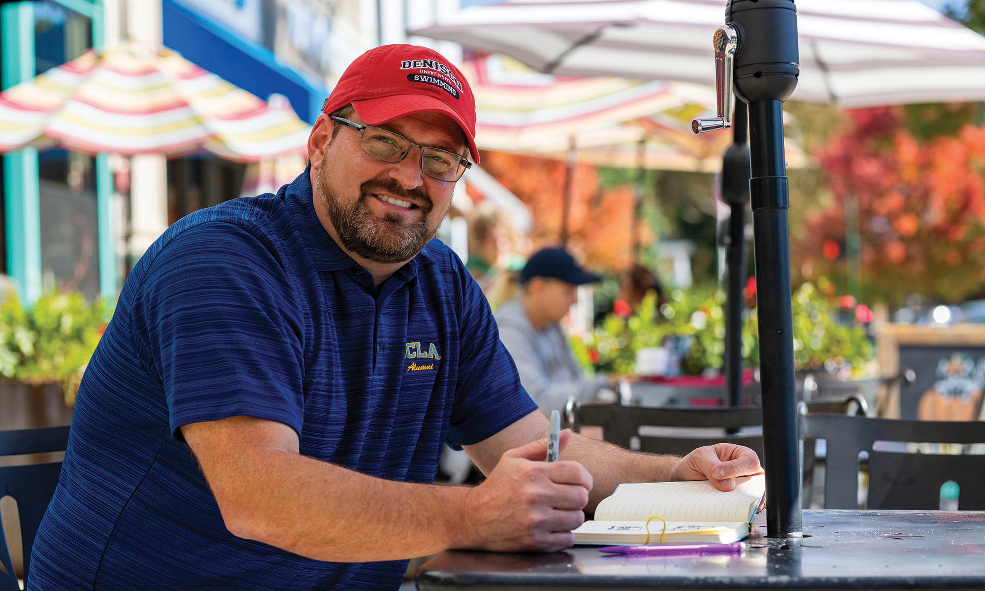 A man in a blue polo shirt and red Denison swimming hat sits at a table on a patio alongside a bustling street.