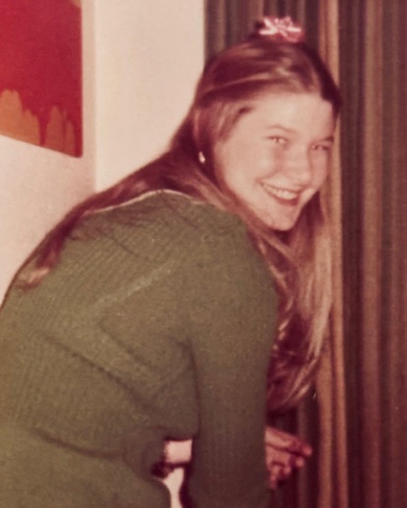 Young woman with long hair wearing a green sweater smiles while leaning forward indoors near a curtain and a wall with artwork.