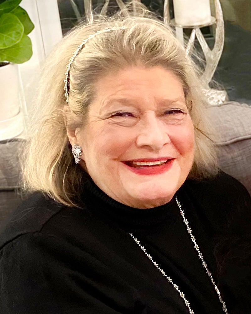 An older woman with light hair, wearing a black top, necklace, and headband, smiles.
