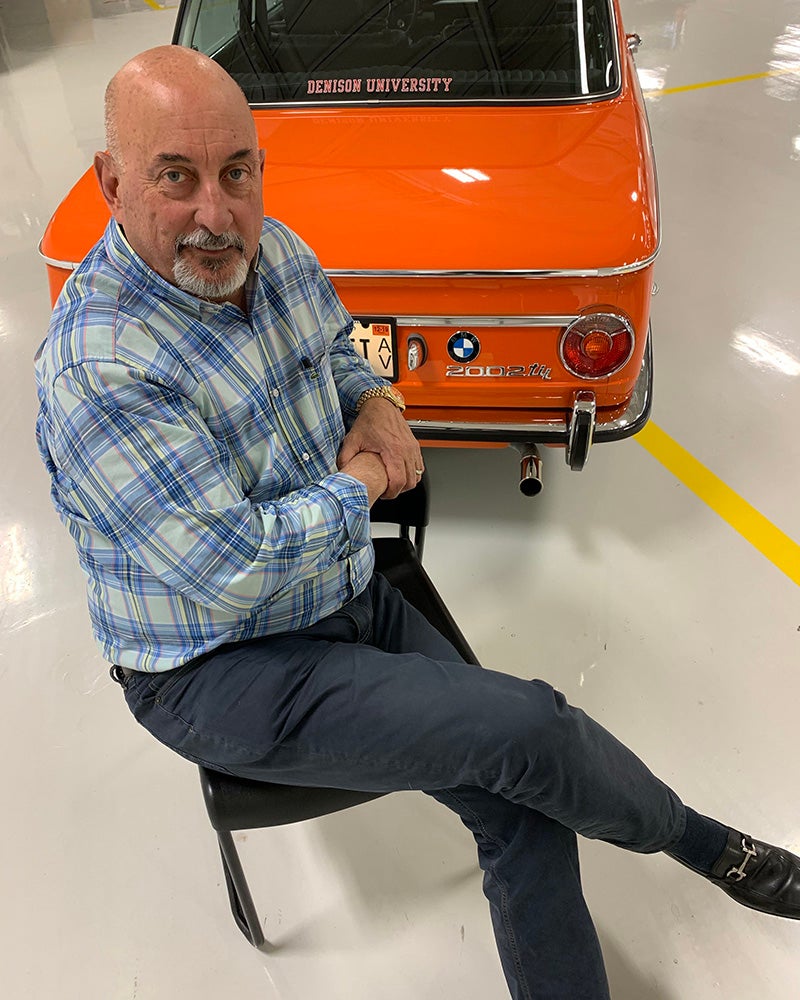 A man in a blue plaid shirt sits on a chair with crossed legs in front of an orange vintage BMW car in a brightly lit garage.