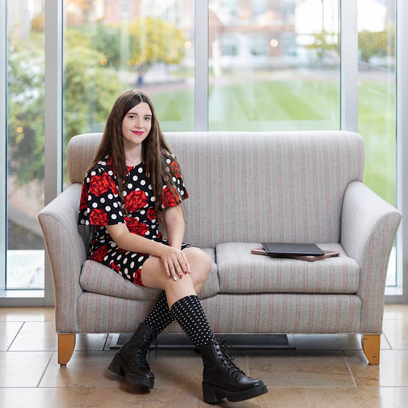 A young woman with long, dark hair, wearing a black dress with a white polka-dot and red flower print, sits on a small, gray couch in front of a large picture window that overlooks a green lawn with trees and buildings in the back ground.