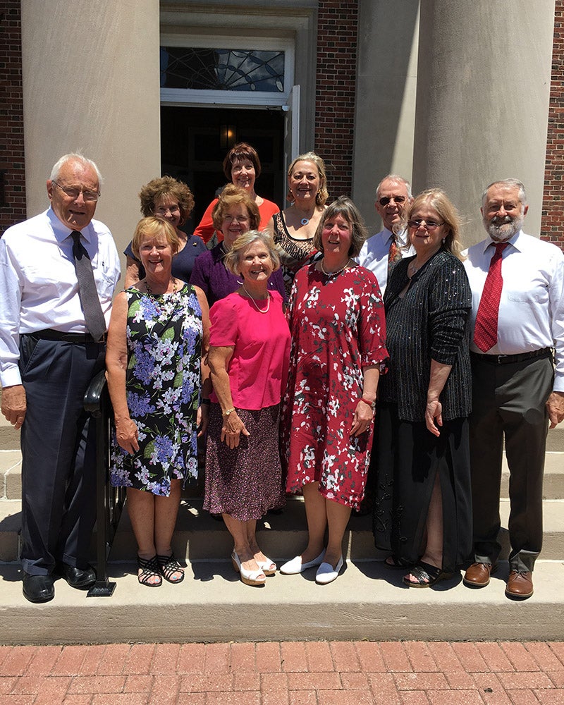A group of eleven adults, both men and women, stand closely together on steps outside a building with large columns, posing for a photo on a sunny day.