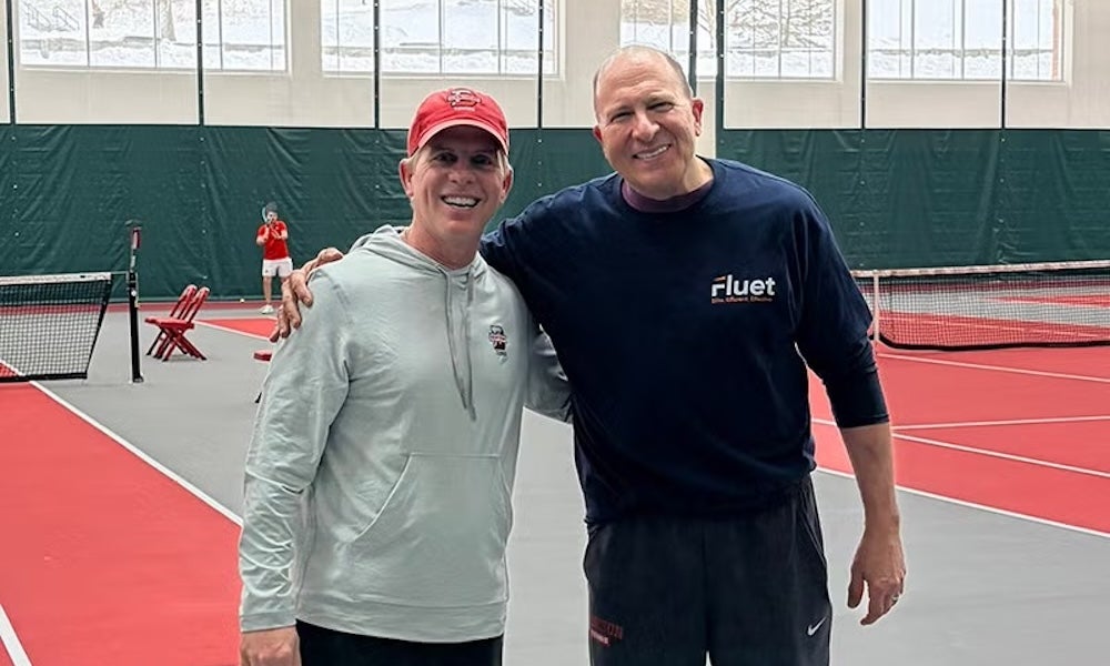 Two men stand side by side on an indoor tennis court. Both are dressed in athletic wear and smiling at the camera. Tennis nets and another person can be seen in the background.