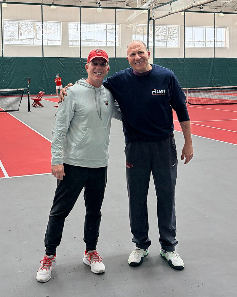 Two men stand side by side on an indoor tennis court. Both are dressed in athletic wear and smiling at the camera. Tennis nets and another person can be seen in the background.