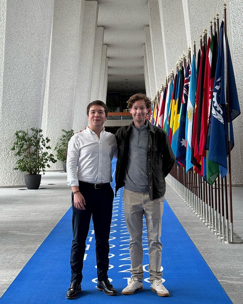 Two young men standing on long blue carpet in a large hallway of columns near a row of international flags.