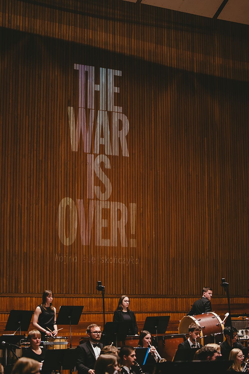 The words "The war is over!" projected in a bold capitalized sanserif font on a brown, wooden bead-board wall above a staged filled with a music ensemble in concert..