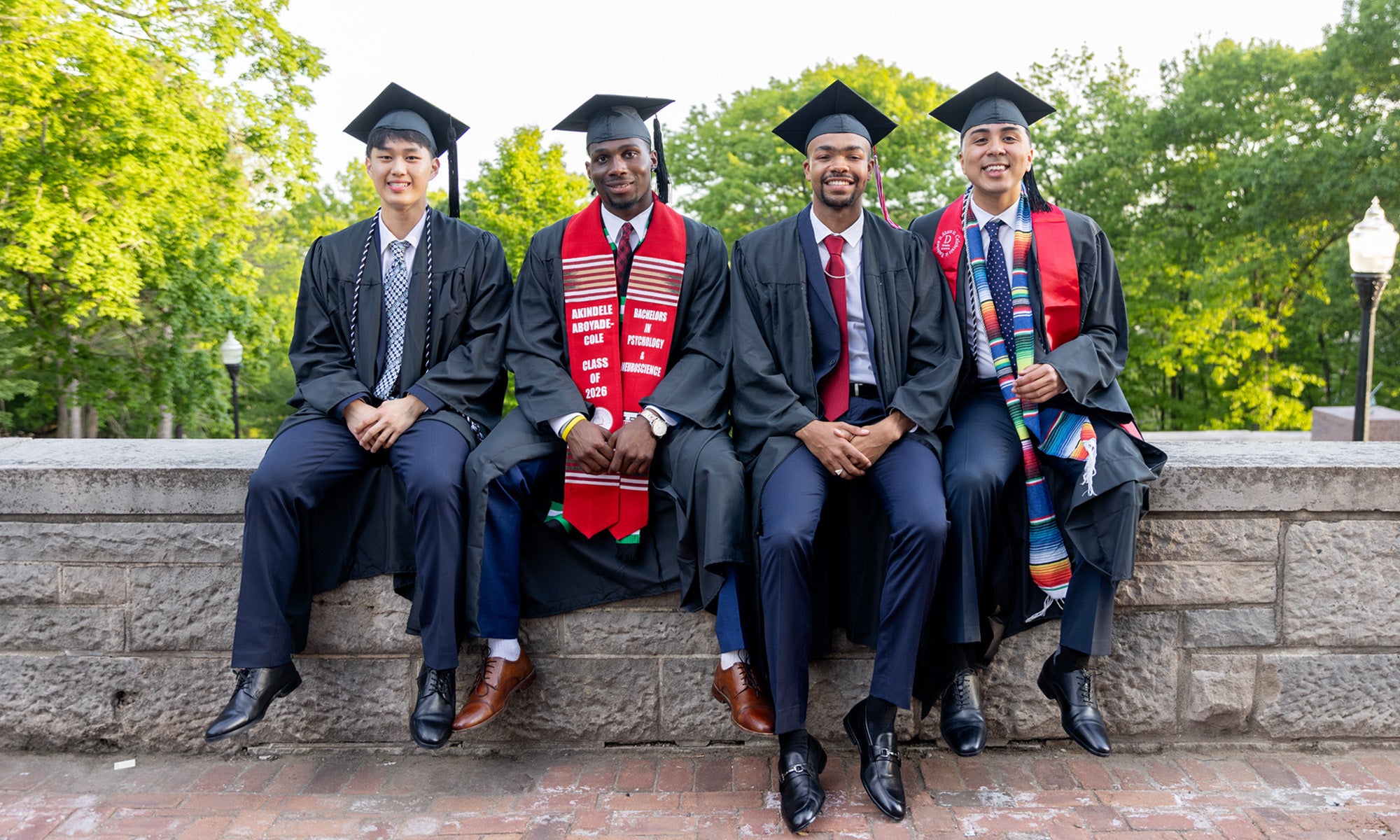 Four young men in graduation caps and gowns sit on a stone wall.