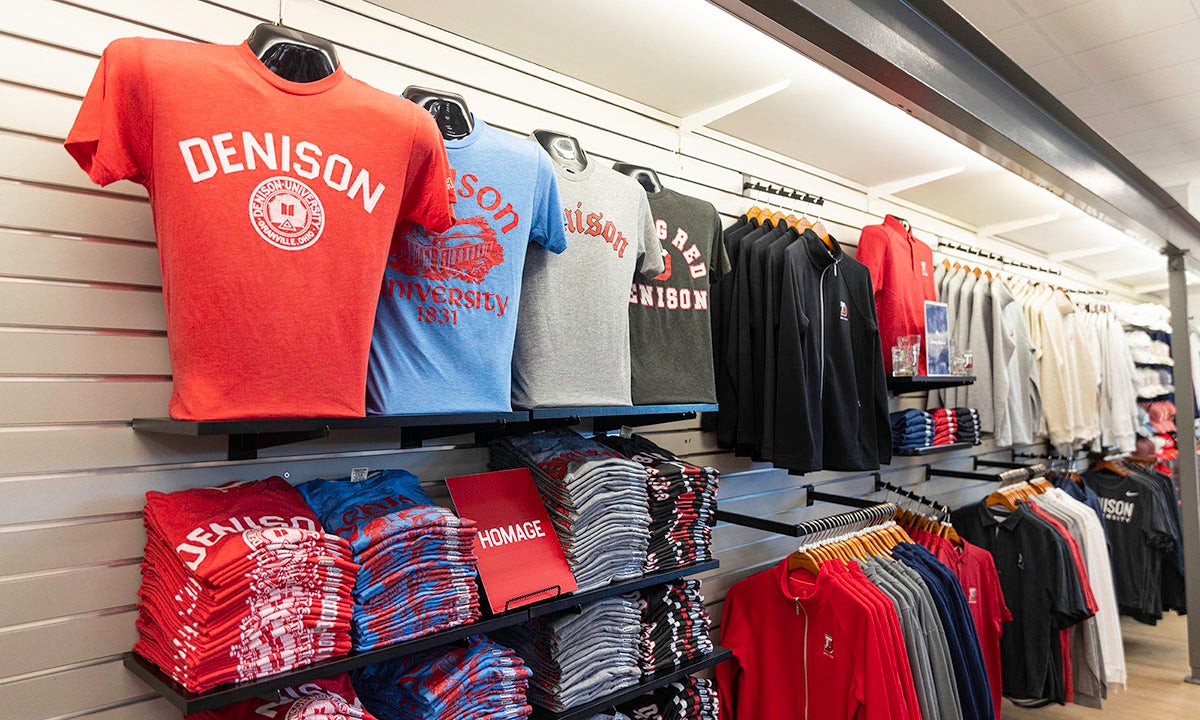 Display of folded and hanging Denison University shirts and sweatshirts in various colors on shelves and racks in a campus bookstore.