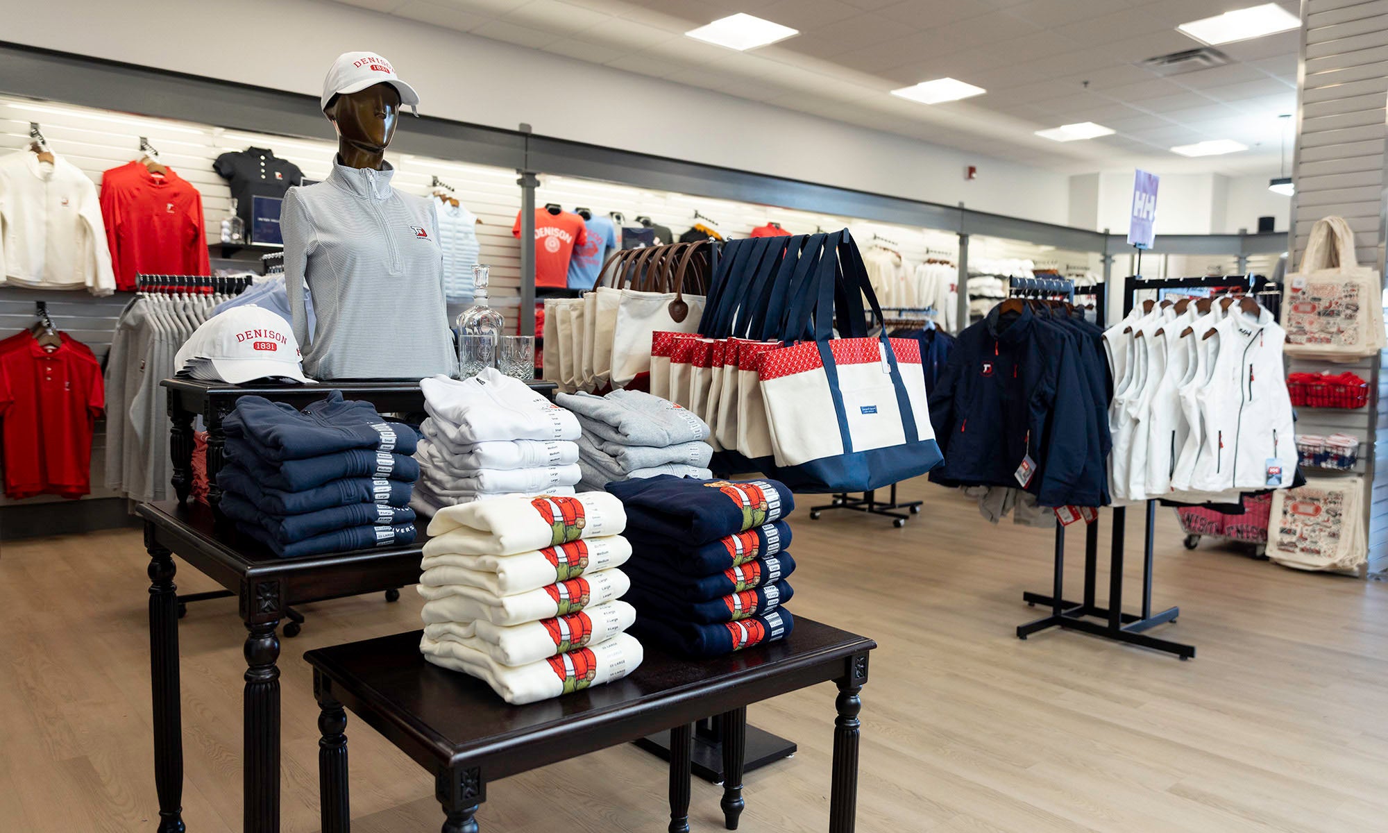 Clothing store interior with folded shirts and sweatshirts on tables, racks of hanging clothes, and tote bags on display.