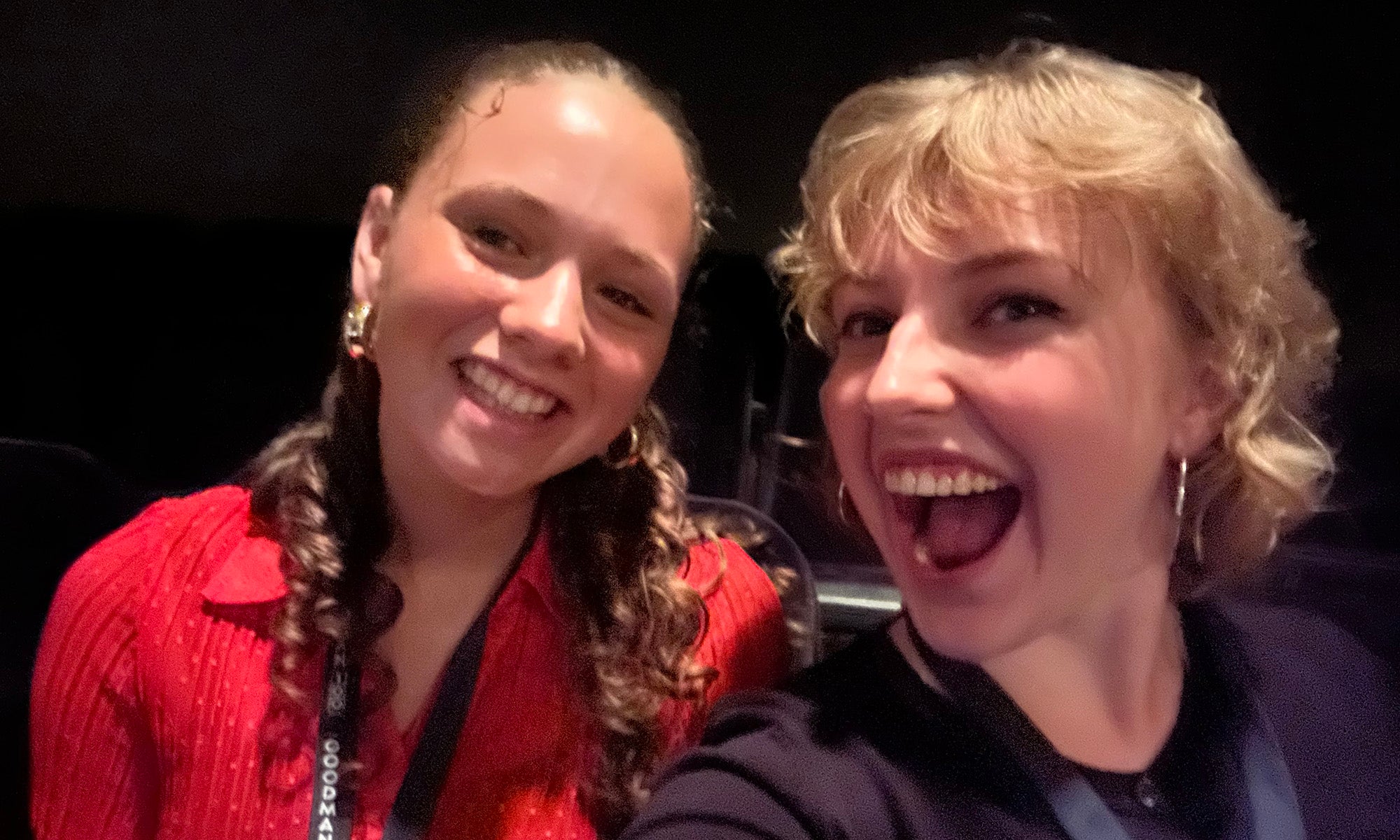Two young women sit together smiling for a selfie in a dimly lit indoor setting.