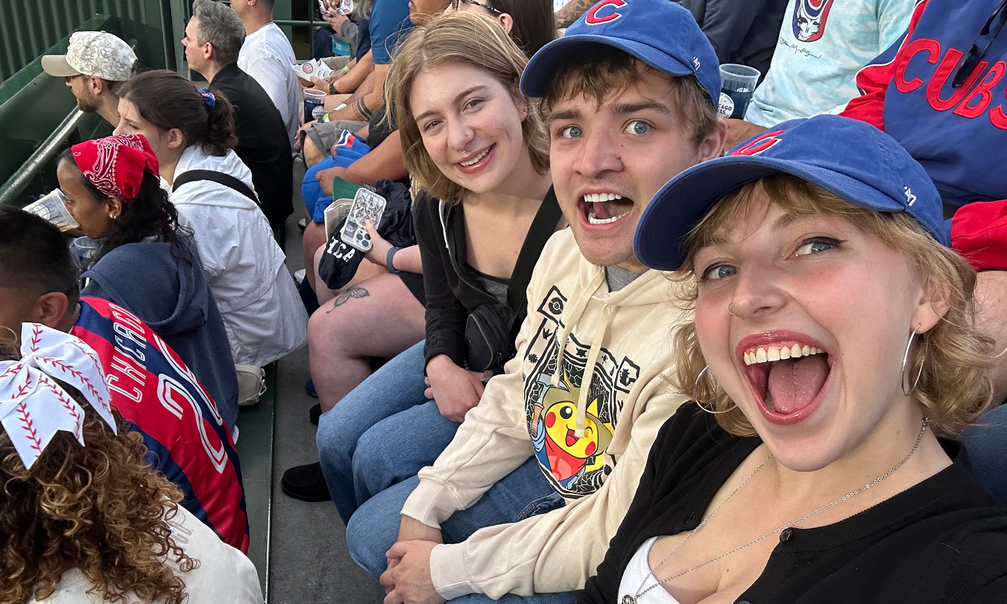 Three young people sit in stadium seats among a crowd, smiling and wearing casual clothing. One person in front wears a blue baseball cap and takes a selfie with the others.