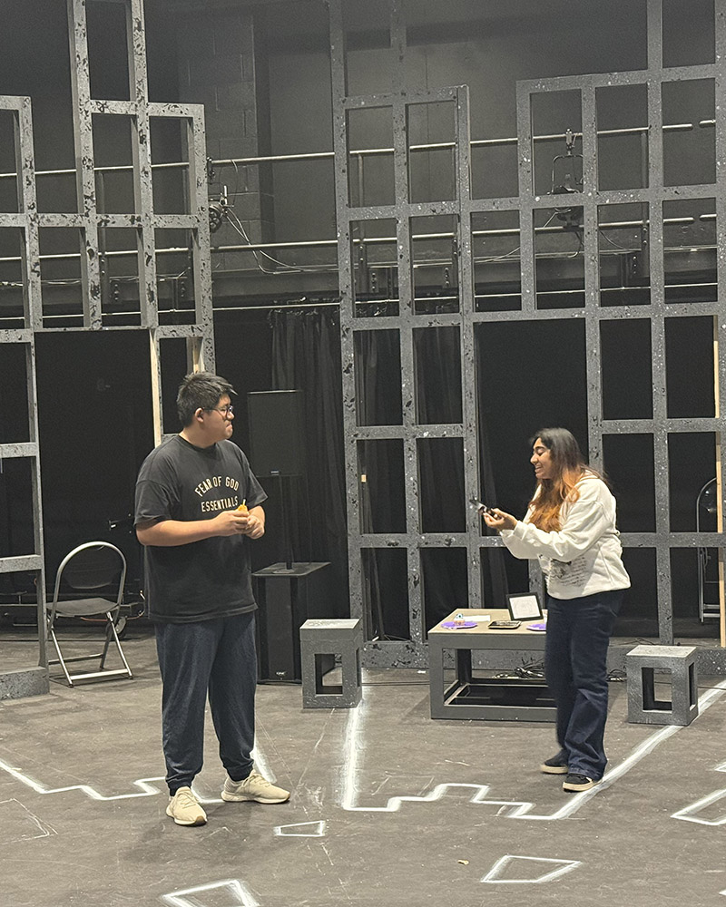 Two people stand on a stage with a geometric set and minimal props. One person holds a phone, while the other faces them. The background features a structure made of large rectangular frames.