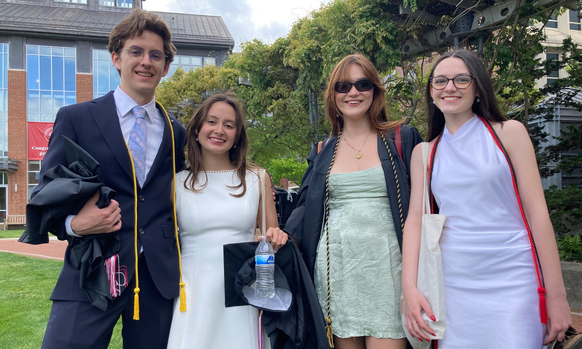 Four students in semi-formal spring wear stand together outside surrounded by greenery.