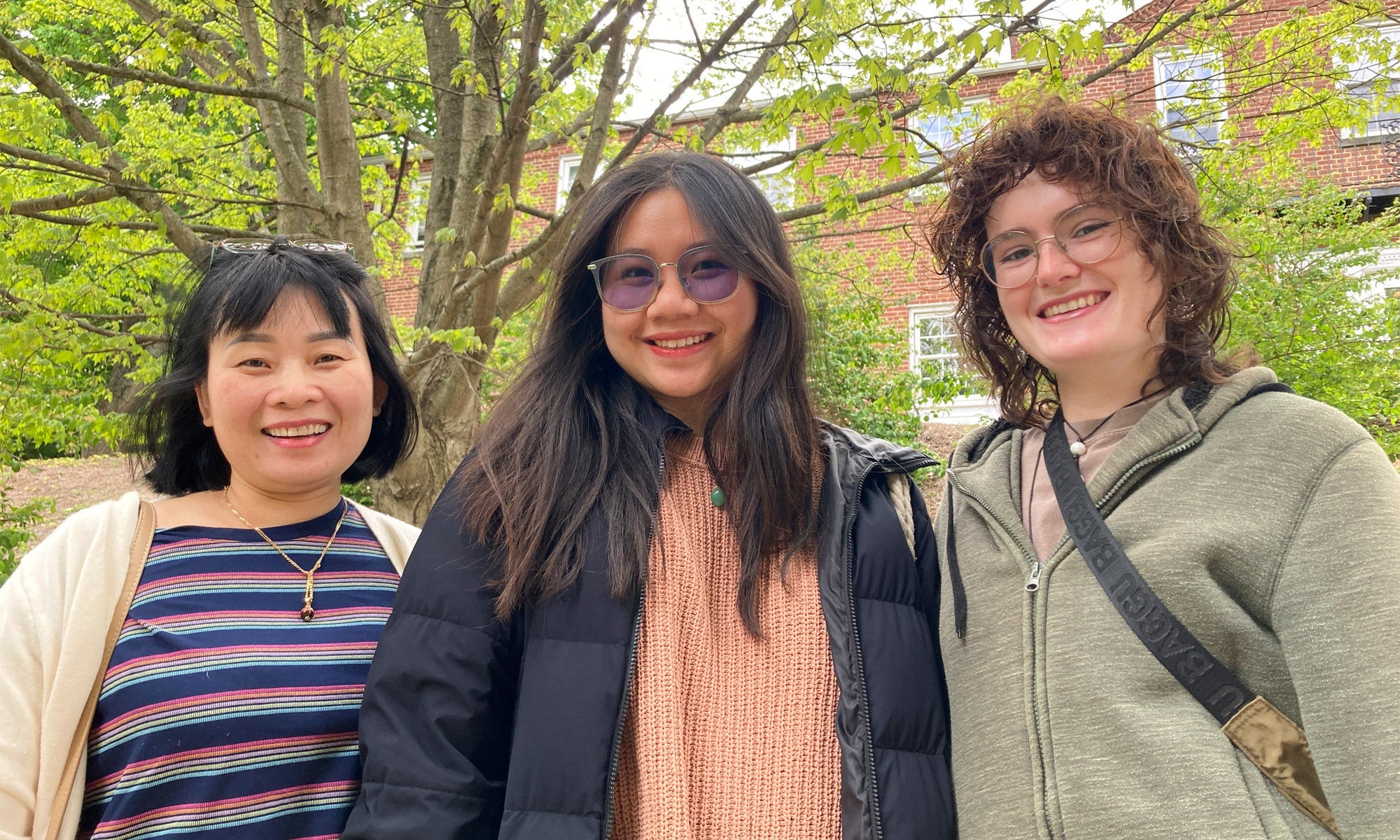 A mother with her college-age daughter and friend pose for a photo outside a brick residence hall.