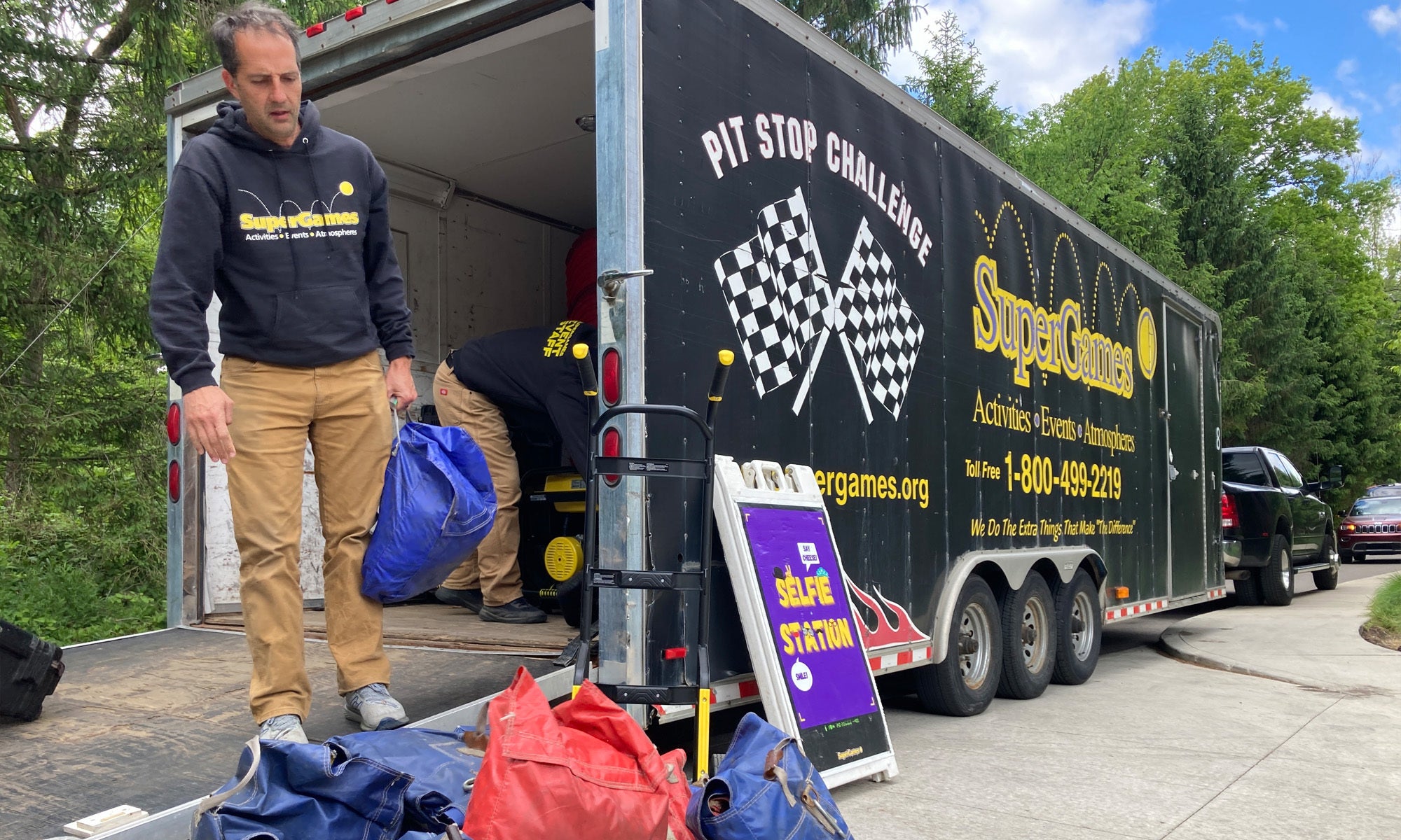 Two men unload bags from a trailer labeled "SuperGames Pit Stop Challenge" parked on a driveway, with equipment and a "SELFIE STATION" sign visible nearby.