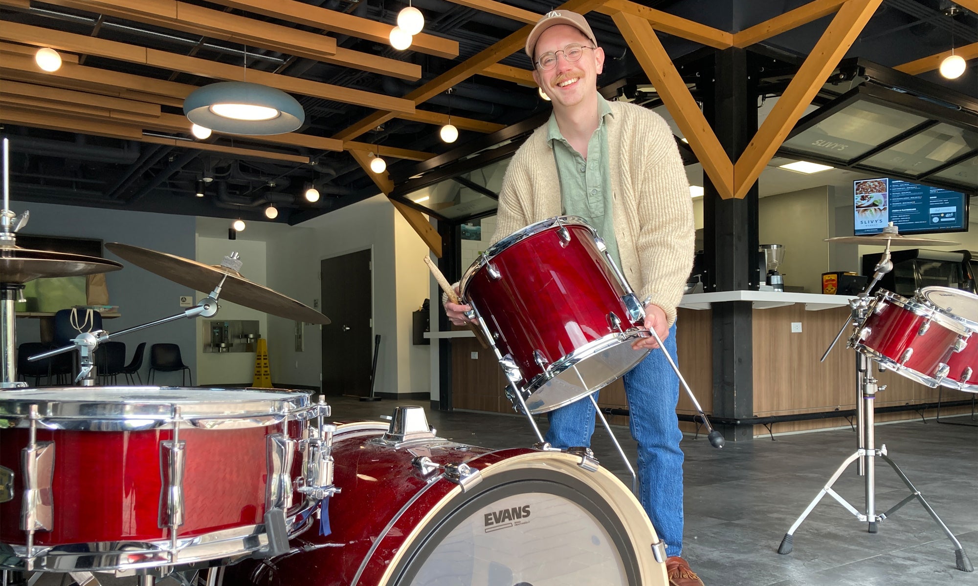 A person wearing glasses and a cardigan holds a red drum in a modern, spacious room with a drum set and wooden ceiling beams.