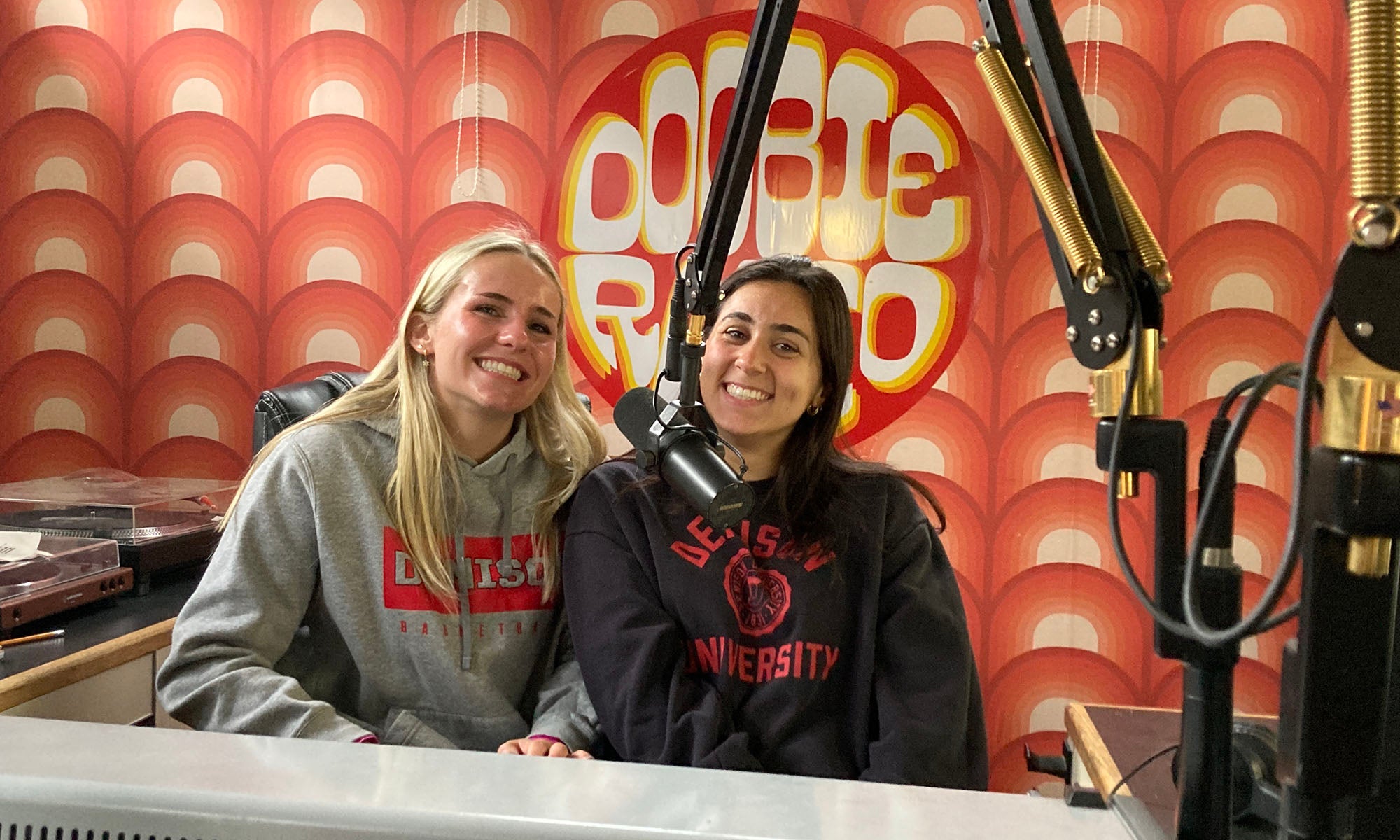 Two young women sit in a radio DJ booth at a microphone; a Doobie Radio sign is on the wall behind them.