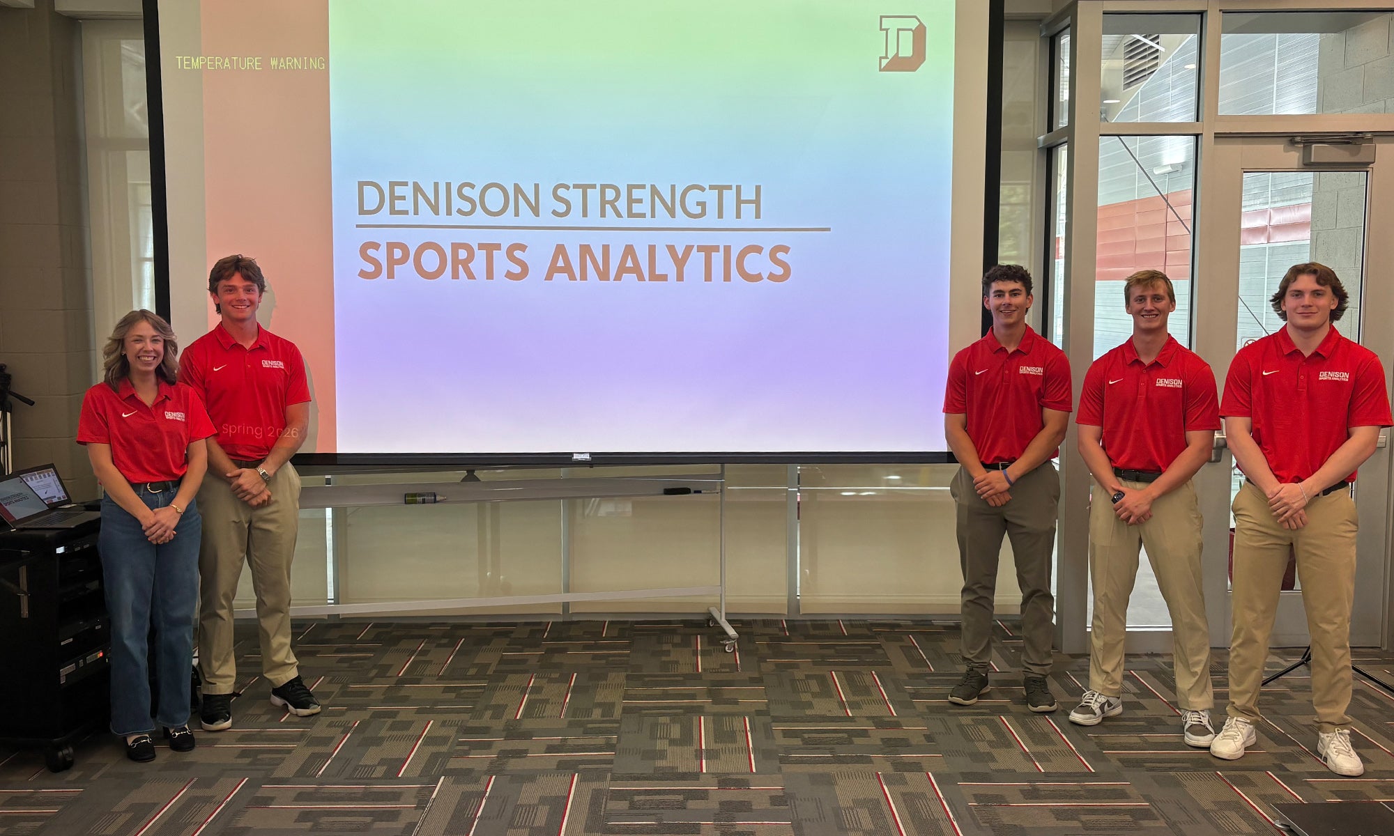 Five people in red shirts and khaki pants stand in front of a projector screen displaying the text Denison Strength Sports Analytics in a classroom or conference room.