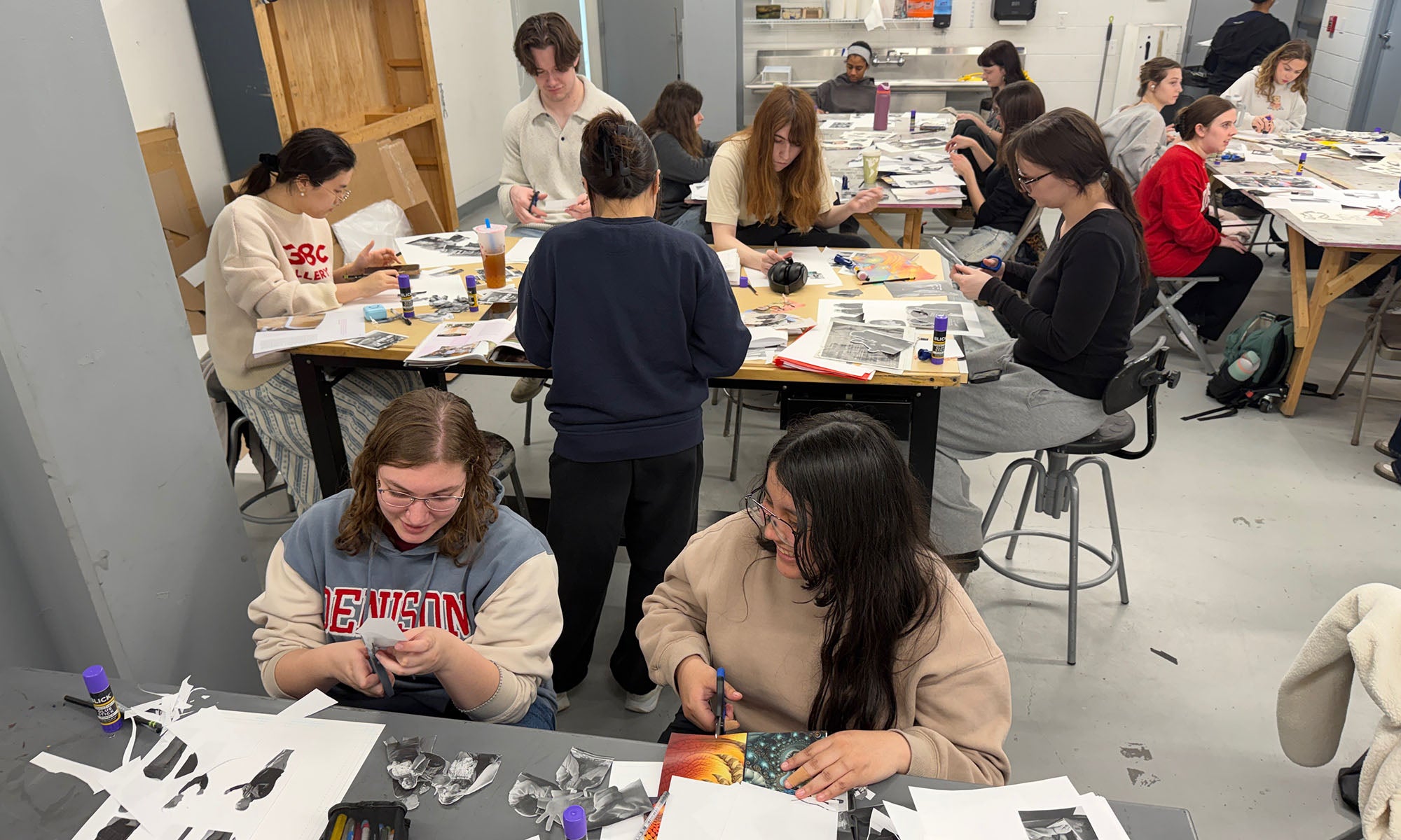 A room of students at tables filled with photos and paper, working on a visual art project.