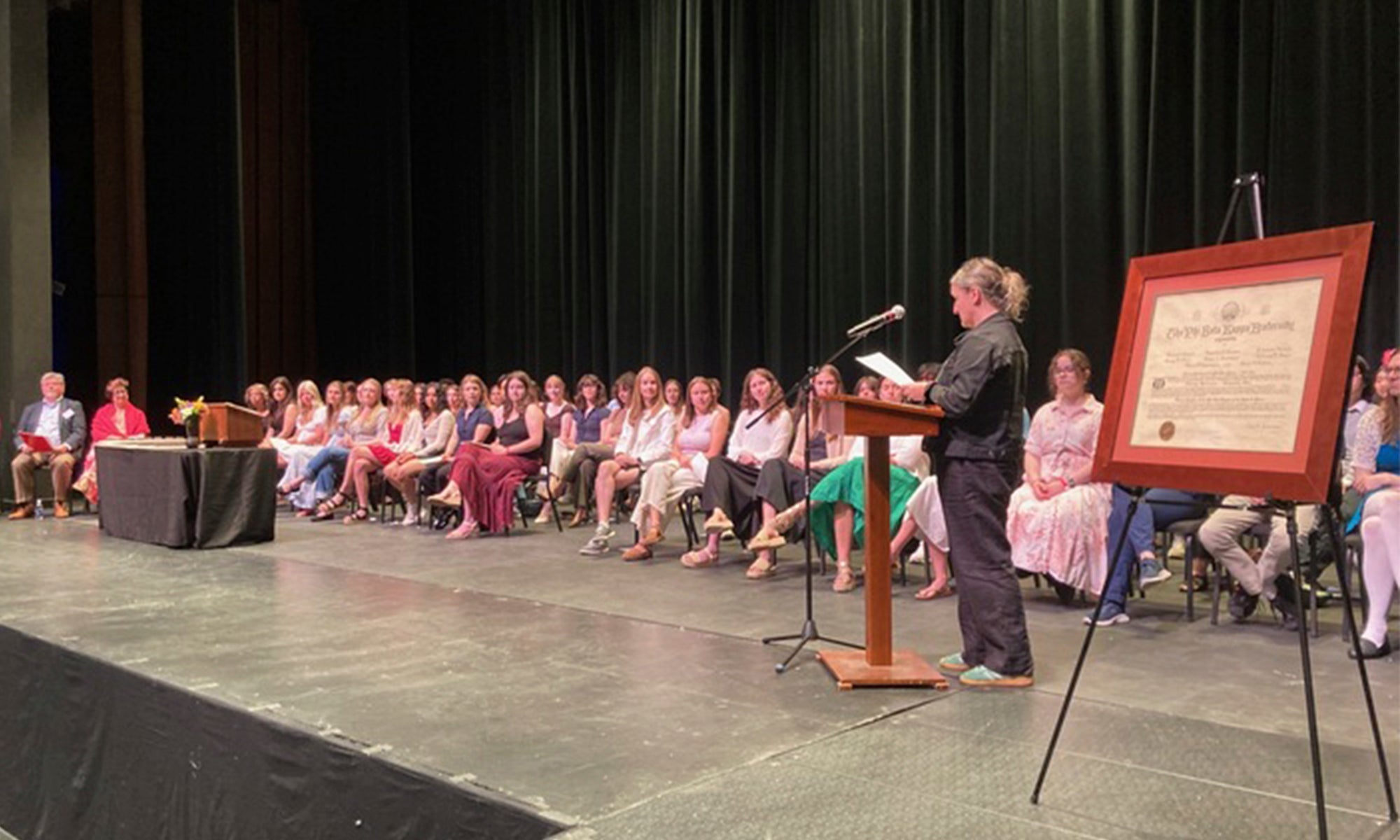 A large group of student on a stage listen to a speaker during a ceremony.