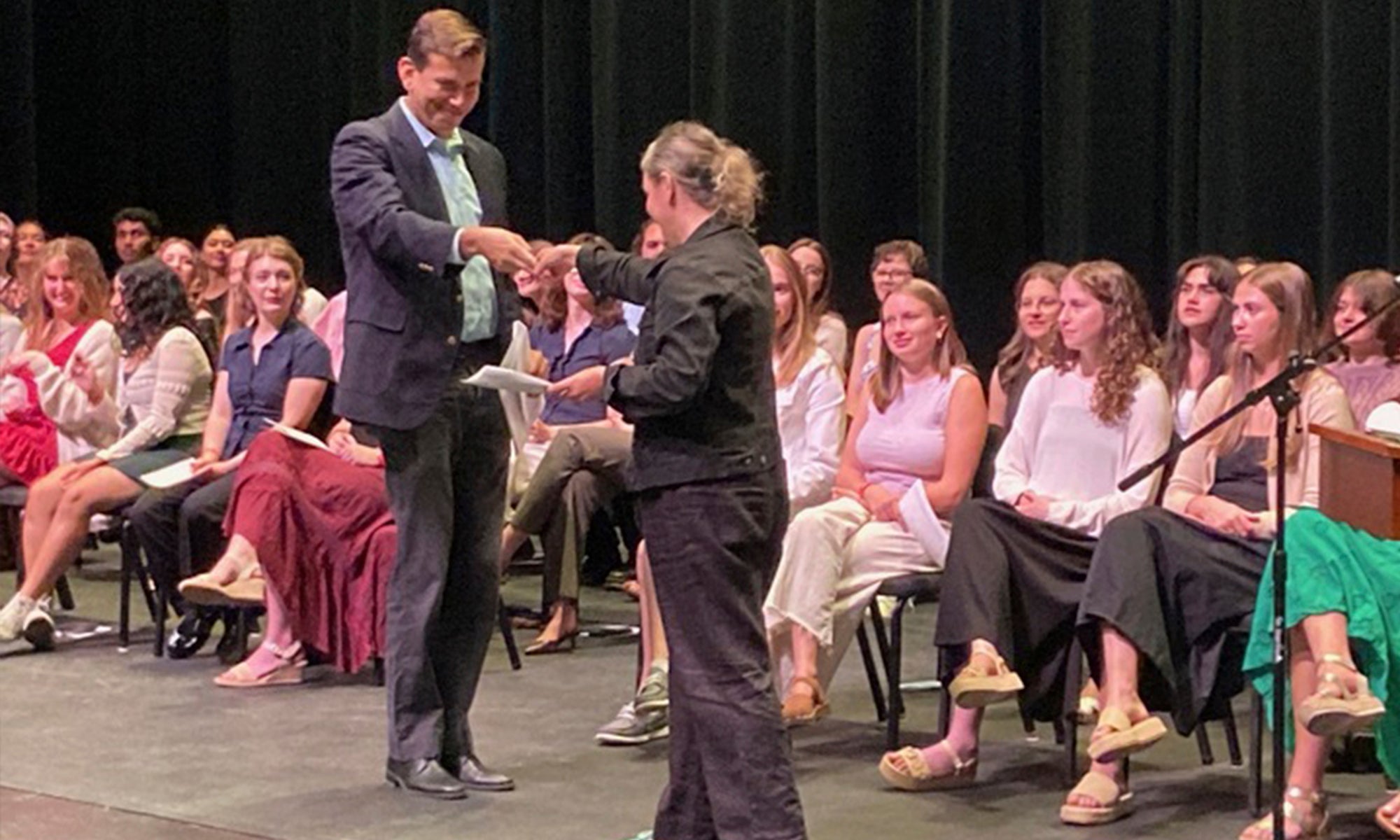 Two people demonstrate a "secret" handshake on stage during a ceremony as a large group of students watch.