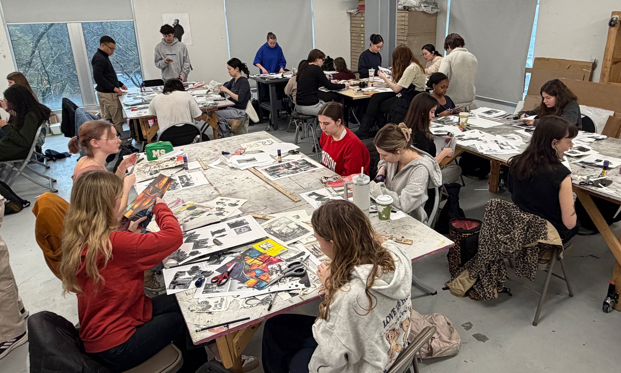 An art room with a dozen or so people sitting at multiple tables that are covered with photos and paper.